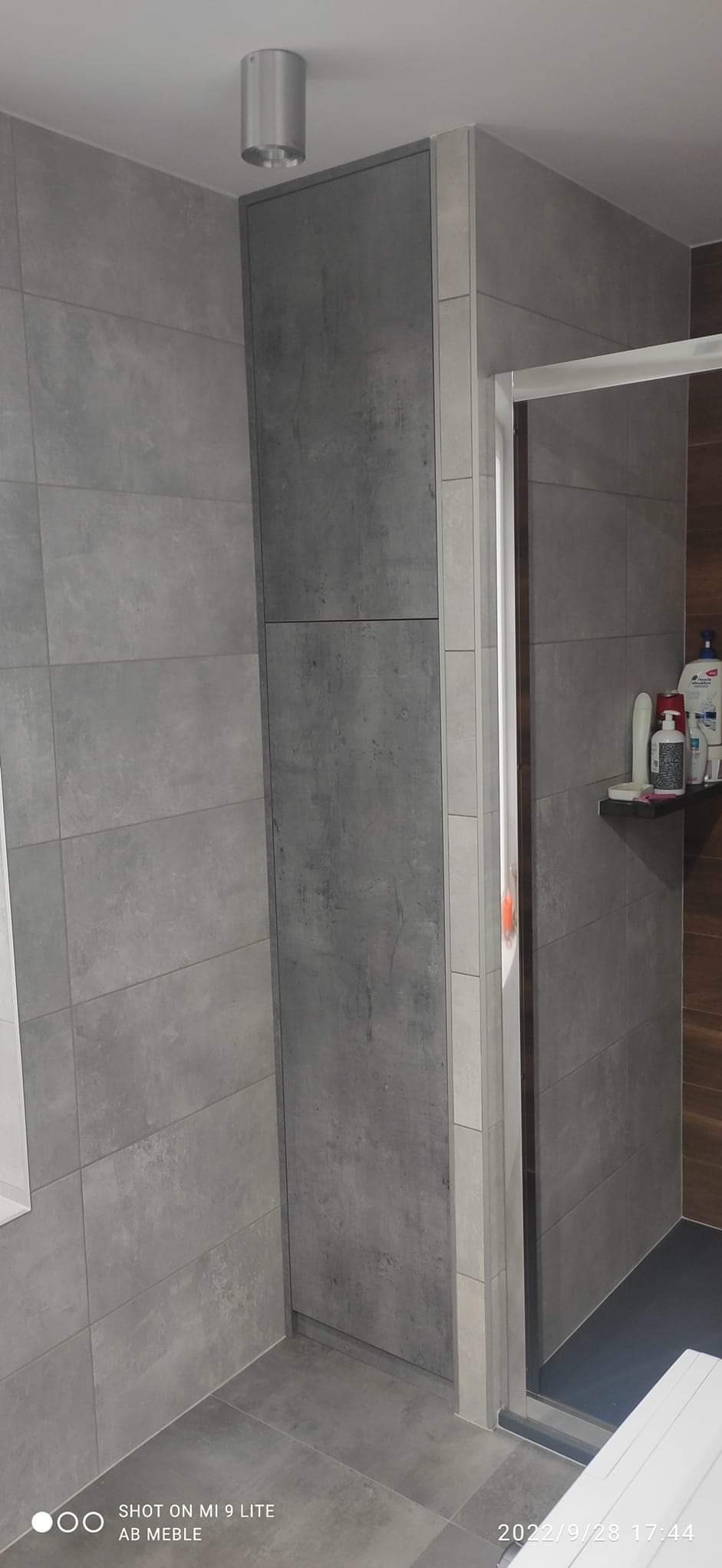 Wysoka, wąska szafka w łazience z szarymi płytkami imitującymi beton, obok kabiny prysznicowej.