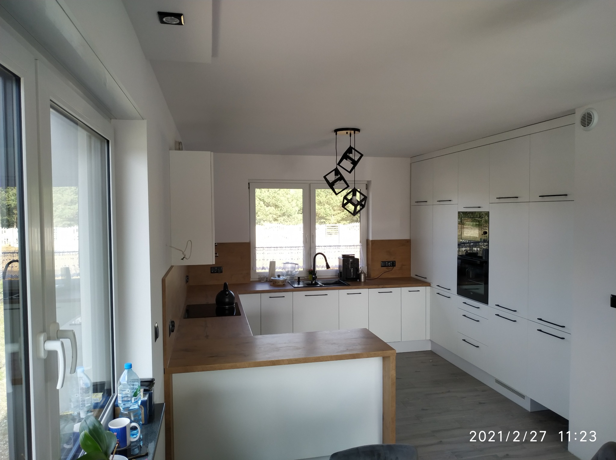 Nowoczesna biała kuchnia z drewnianym blatem i wyspą, oświetlona geometryczną lampą wiszącą. Widok na okno z zielenią w tle.