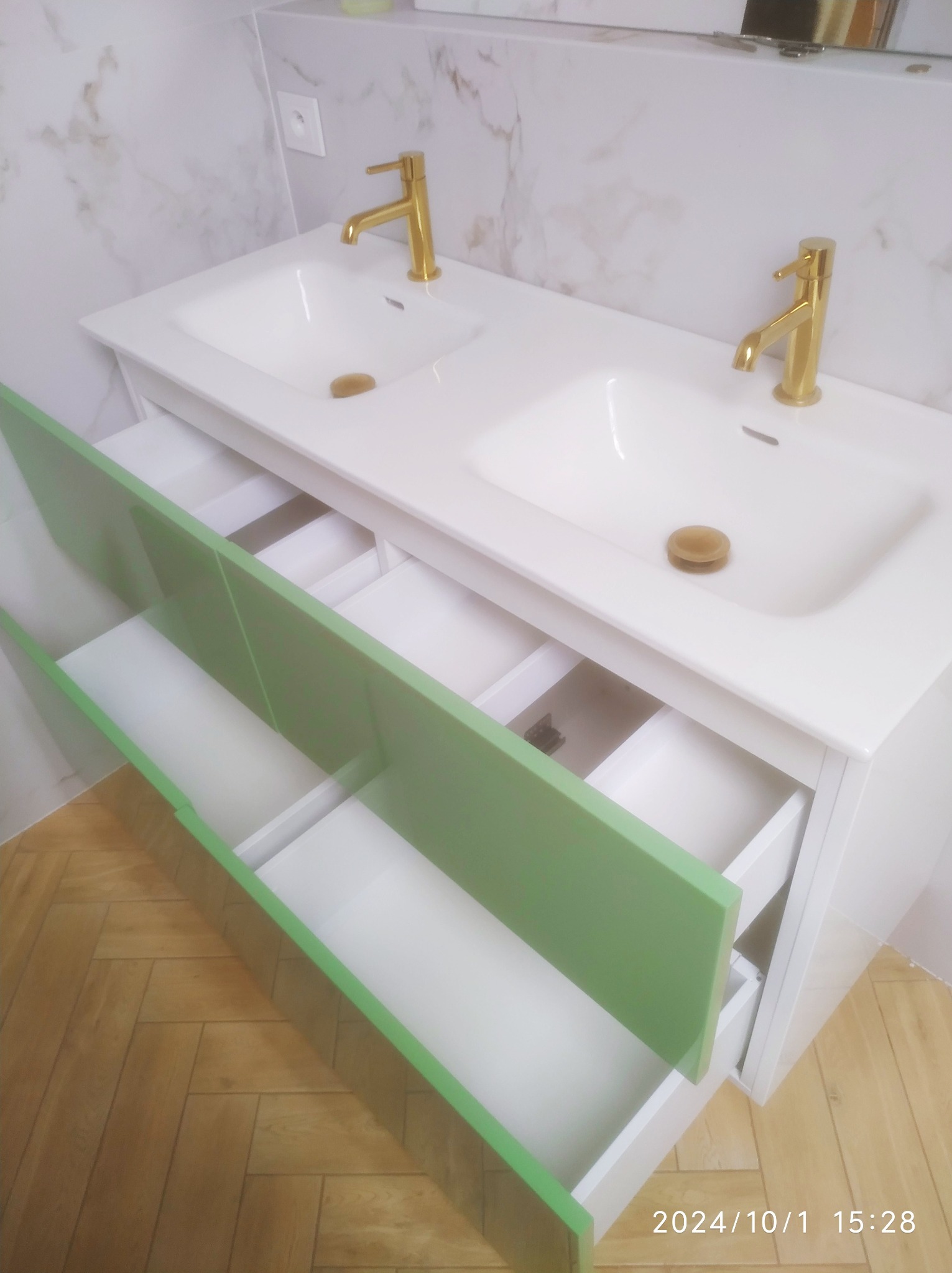 Biała umywalka podwójna z otwartymi szufladami w kolorze jasnej zieleni i złotą armaturą na tle marmurowej ściany i parkietu ułożonego w jodełkę.