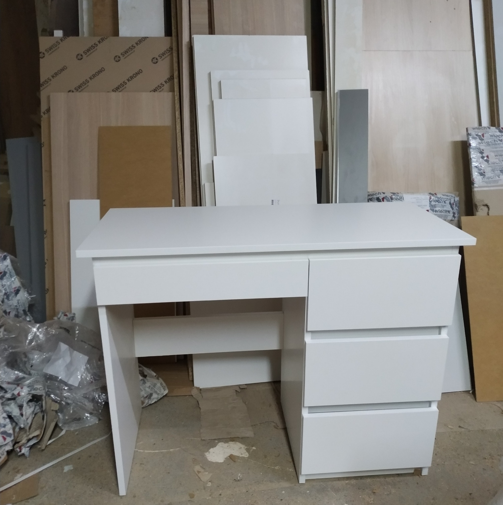 Białe biurko z szufladami ustawione w warsztacie stolarskim, w tle płyty meblowe Swiss Krono i opakowania folii.