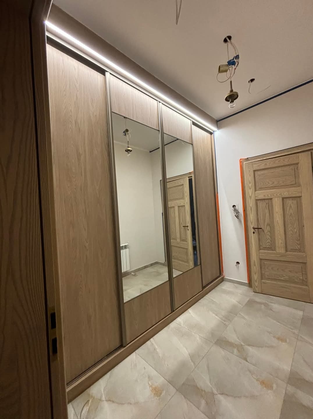 Szafa wnękowa z przesuwnymi drzwiami z lustrem, oświetlenie LED, drewniane wykończenie, widoczne niedokończone prace elektryczne sufitowe i listwy przypodłogowe.