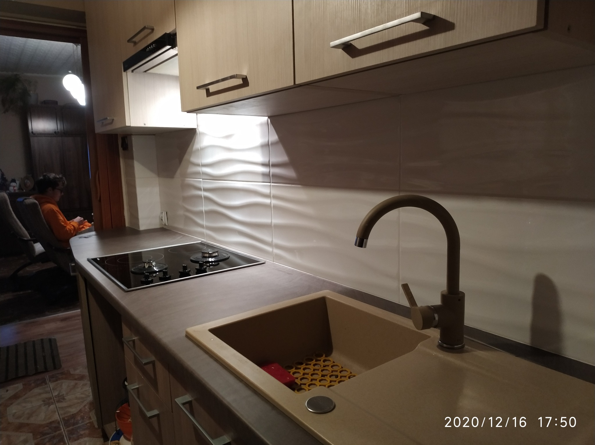 Beżowa kuchnia z drewnianymi szafkami, płytą indukcyjną i zlewozmywakiem z brązową baterią, widok na salon z osobą siedzącą w fotelu.