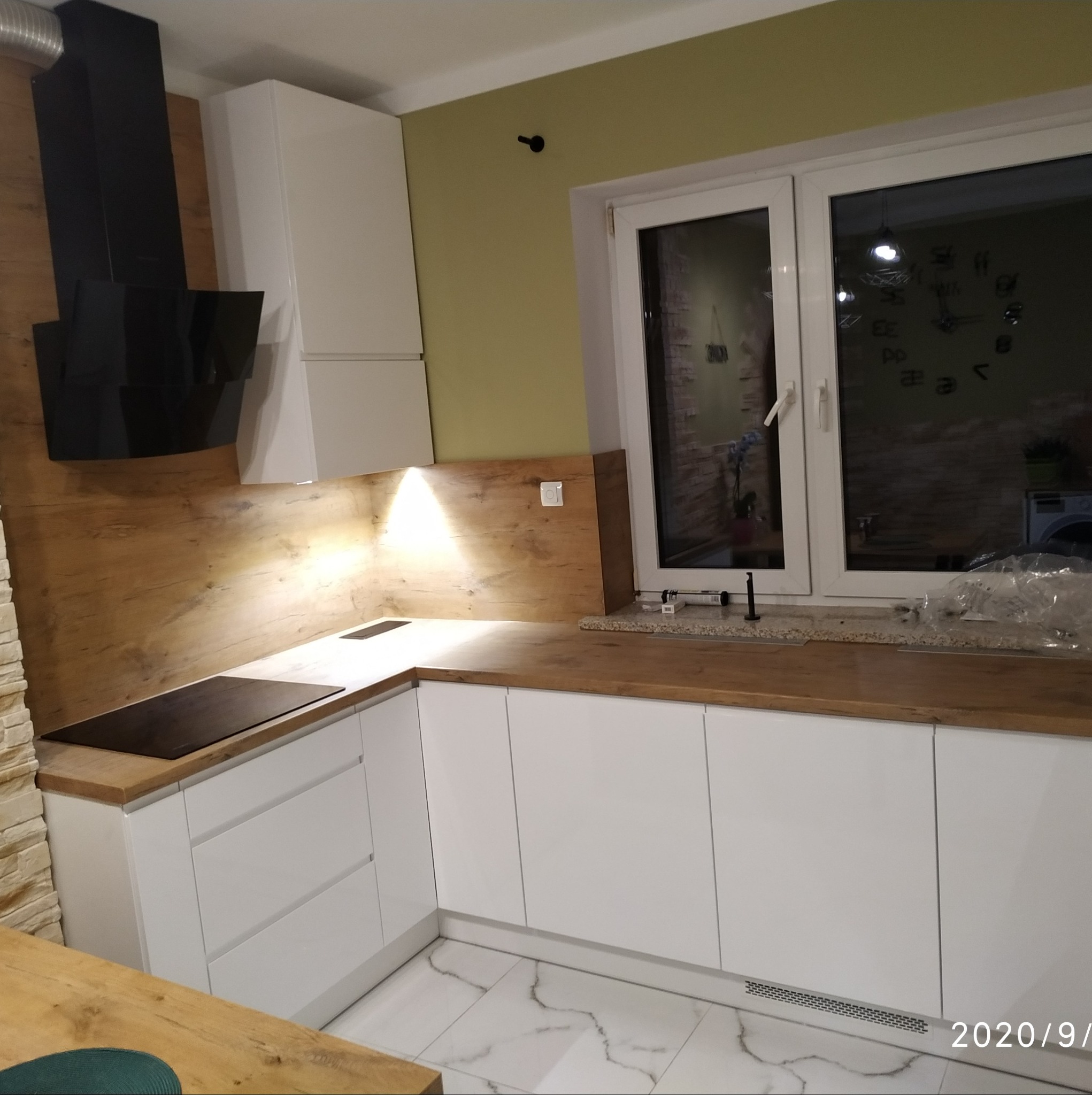 Nowoczesna kuchnia z białymi frontami szafek, drewnianym blatem i okładziną ścienną, czarnym okapem oraz płytą indukcyjną. W tle widoczne okno z odbiciem zegara na ścianie.