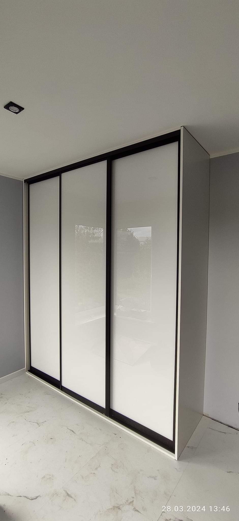 Nowoczesna szafa wnękowa z trzema przesuwnymi, białymi frontami z czarnymi obramowaniami, widoczna na tle szarej ściany i marmurowej podłogi.