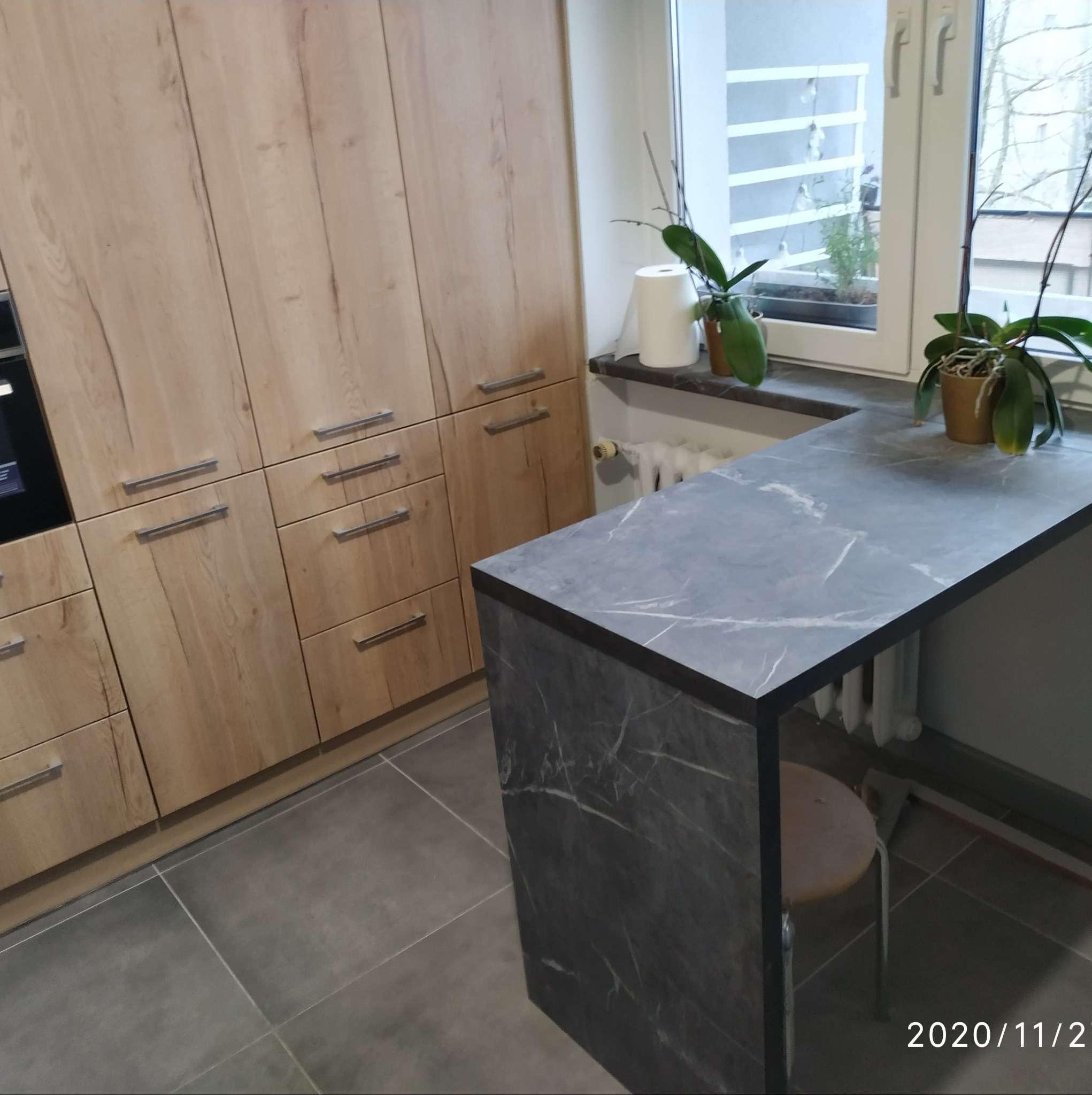 Nowoczesna kuchnia z drewnianymi szafkami, szarym blatem imitującym marmur i małym stołkiem barowym, z widokiem na rośliny doniczkowe na parapecie.