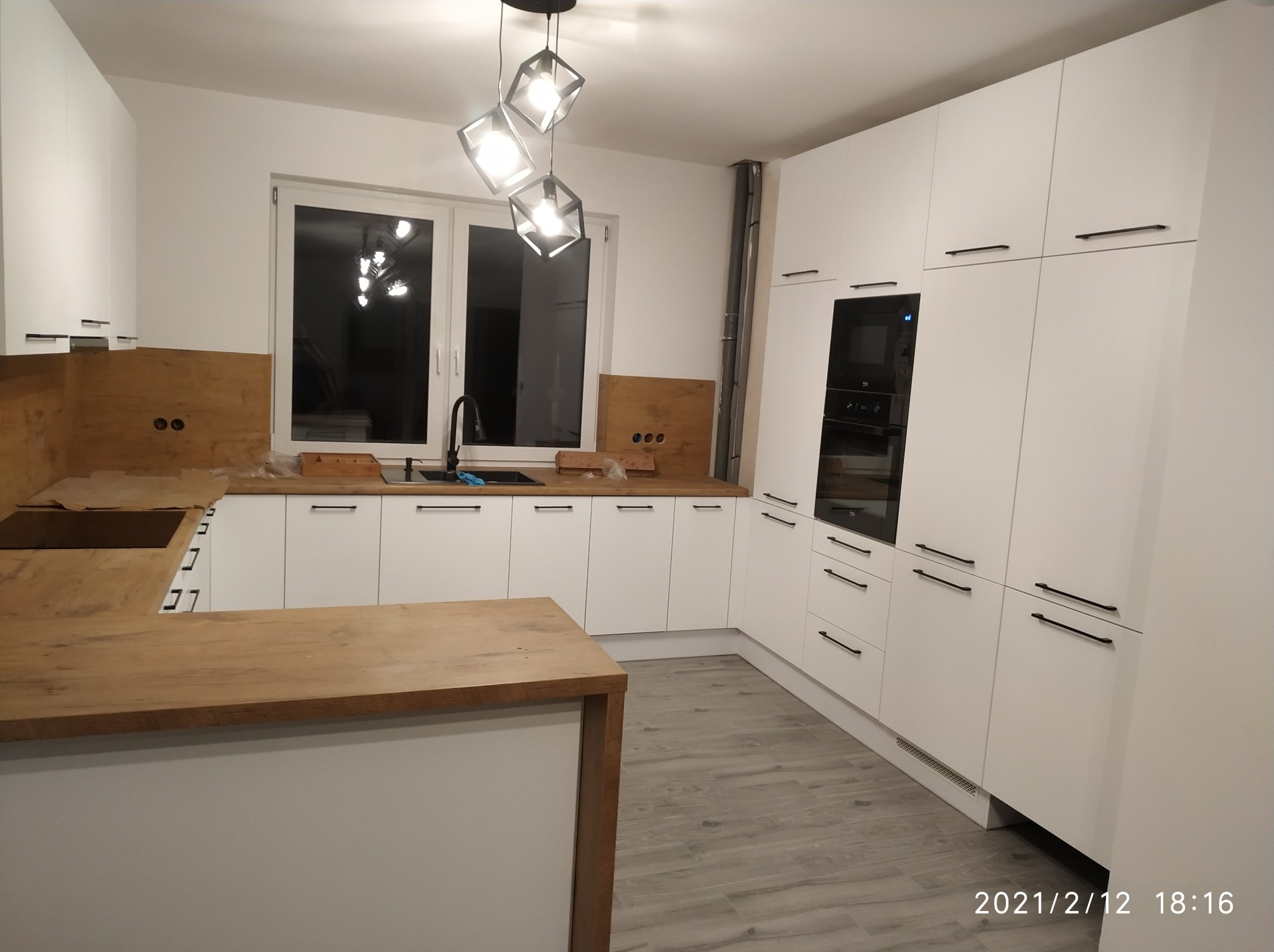 Nowoczesna kuchnia z białymi szafkami, drewnianym blatem i wyspą kuchenną, oświetlona geometryczną lampą wiszącą. Widoczne okno i fragment instalacji wentylacyjnej.