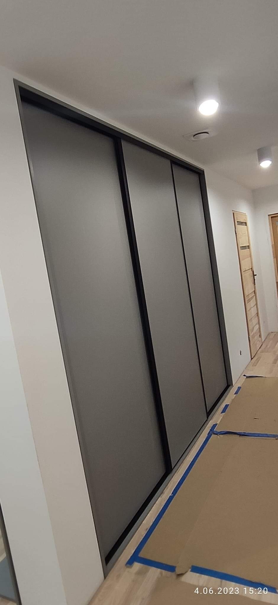 Zamknięta szafa wnękowa z przesuwnymi drzwiami w czarnej ramie, zabezpieczona kartonem i taśmą malarską na podłodze