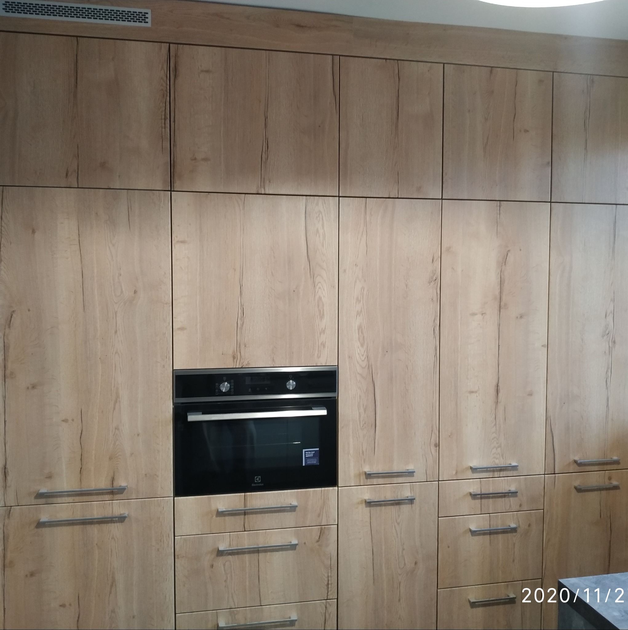 Jasna zabudowa kuchenna z drewnopodobnymi frontami, z wbudowanym piekarnikiem Electrolux i metalowymi uchwytami.