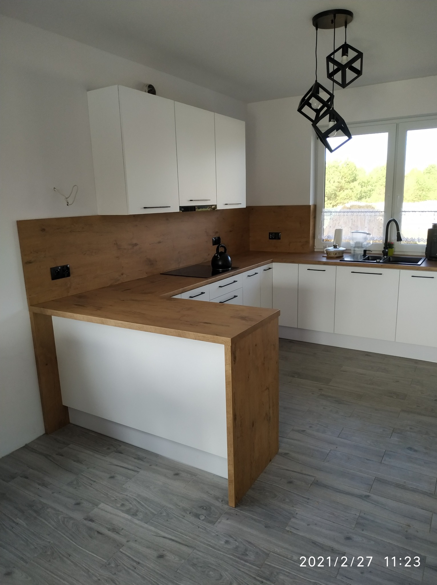 Nowoczesna kuchnia z białymi szafkami i drewnianym blatem, czarny czajnik na płycie indukcyjnej, geometryczna lampa wisząca, szare panele podłogowe.