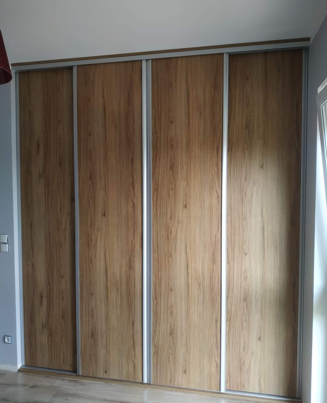 Czterodrzwiowa szafa przesuwna z frontami w okleinie imitującej jasne drewno, z aluminiowymi ramami, zamontowana w pokoju z szarymi ścianami.