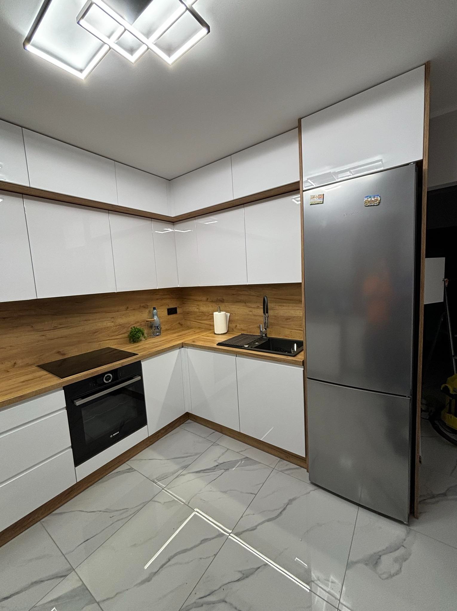 Nowoczesna kuchnia z białymi, lakierowanymi frontami, drewnianym blatem i szarą lodówką Bosch, oświetlona geometryczną lampą sufitową, uzupełniona czarnym zlewem i płytą indukcyjną.