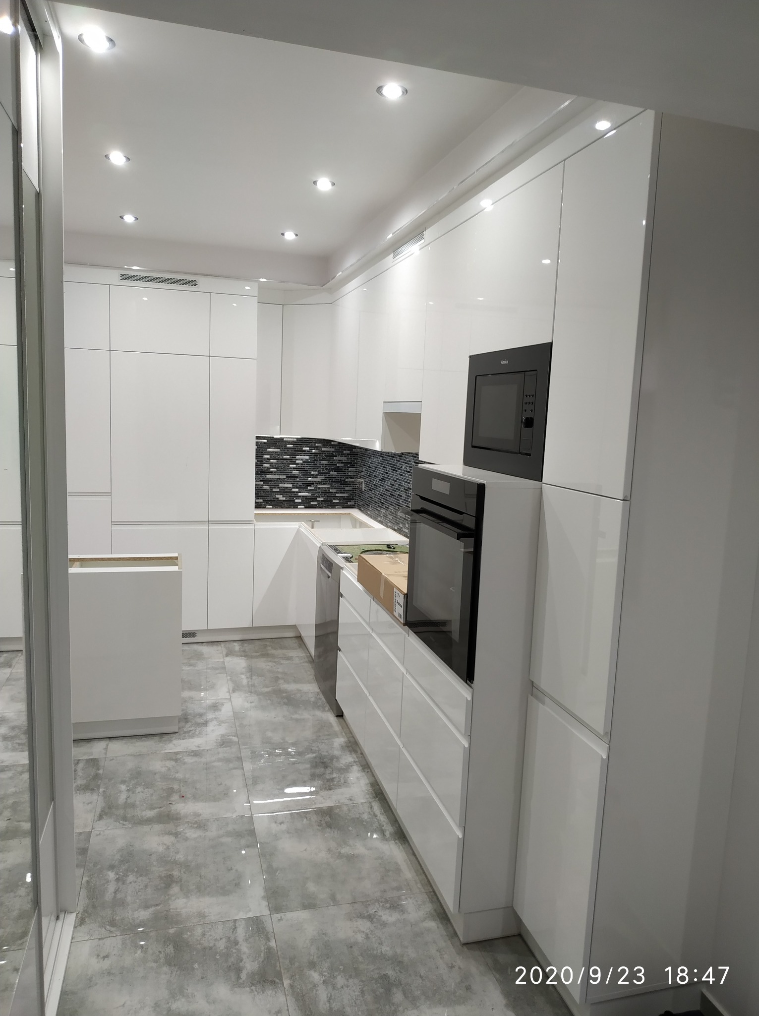 Nowoczesna, biała kuchnia z błyszczącymi frontami, czarną mozaiką nad blatem i wbudowanym sprzętem AGD, w trakcie montażu.