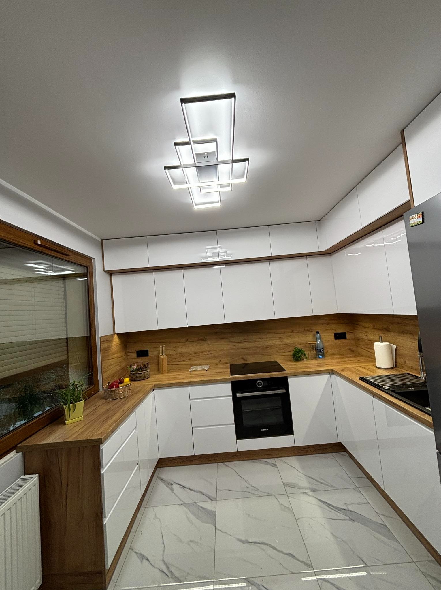 Nowoczesna kuchnia z białymi frontami i drewnianym blatem, oświetlona geometryczną lampą sufitową, widok na fragment okna i marmurową podłogę.
