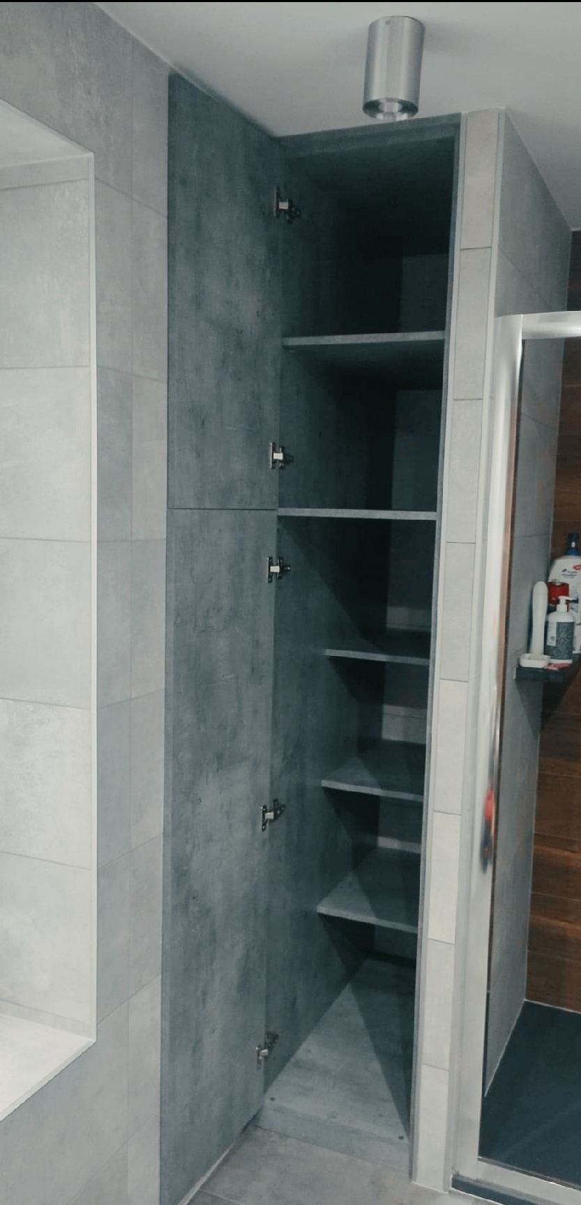 Wysoka, wąska szafa wnękowa z otwartymi drzwiami, wykończona szarą okładziną imitującą beton, z półkami wewnątrz, zamontowana w łazience wyłożonej jasnymi płytkami.