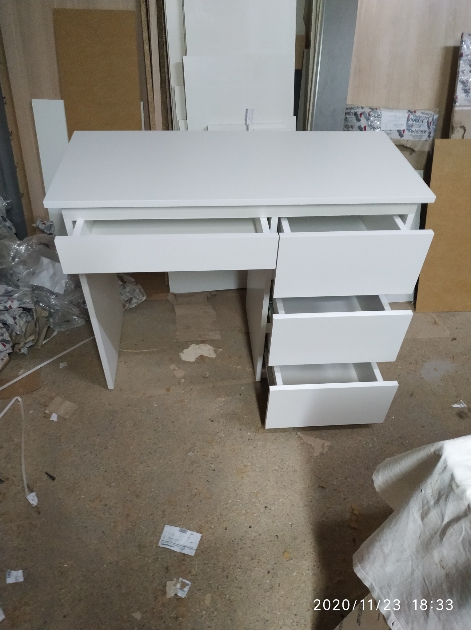 Białe biurko z szufladami w warsztacie stolarskim, jedna szuflada wysunięta, widoczne płyty meblowe w tle i data na podłodze.