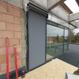 Montaż szarej rolety antywłamaniowej zewnętrznej przy oknie aluminiowym, widoczne poziomice i wkrętarka na tle surowej ściany z cegły i nie wykończonego dachu.