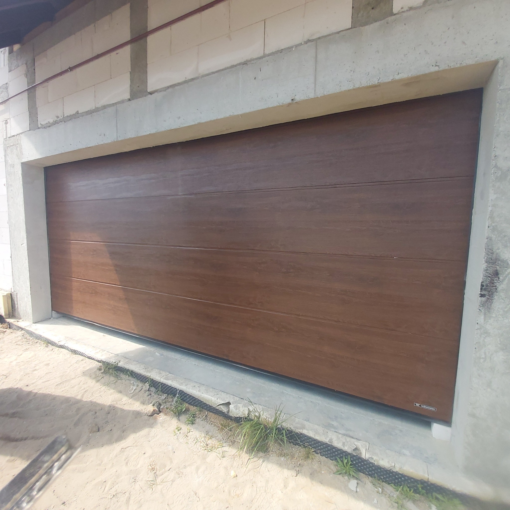 Zamknięta, segmentowa brama garażowa w kolorze drewna, zamontowana w surowym otworze budynku w trakcie budowy, widoczne bloczki betonowe i piasek.