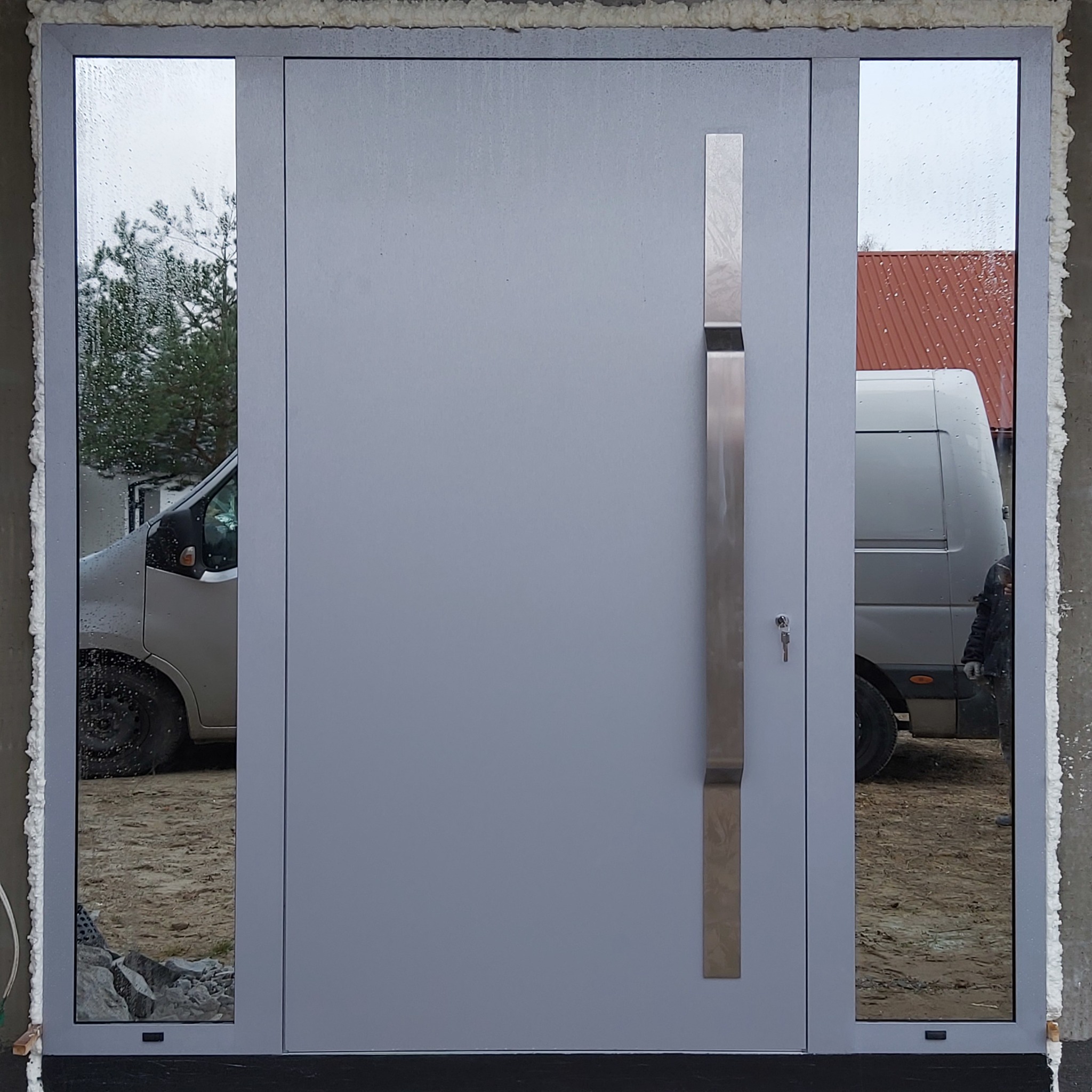 Nowoczesne, szare drzwi zewnętrzne z długim, pionowym uchwytem i bocznymi szybami, zamontowane z widoczną pianką montażową, odbijające w szybach samochody dostawcze.