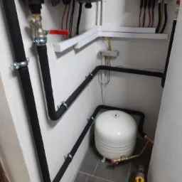 Instalacja z białym zbiornikiem wyrównawczym, czarnymi rurami izolowanymi i pompą obiegową, widoczne okablowanie elektryczne i skrzynka bezpiecznikowa na ścianie, w tle płytki podłogowe, w pobliżu...