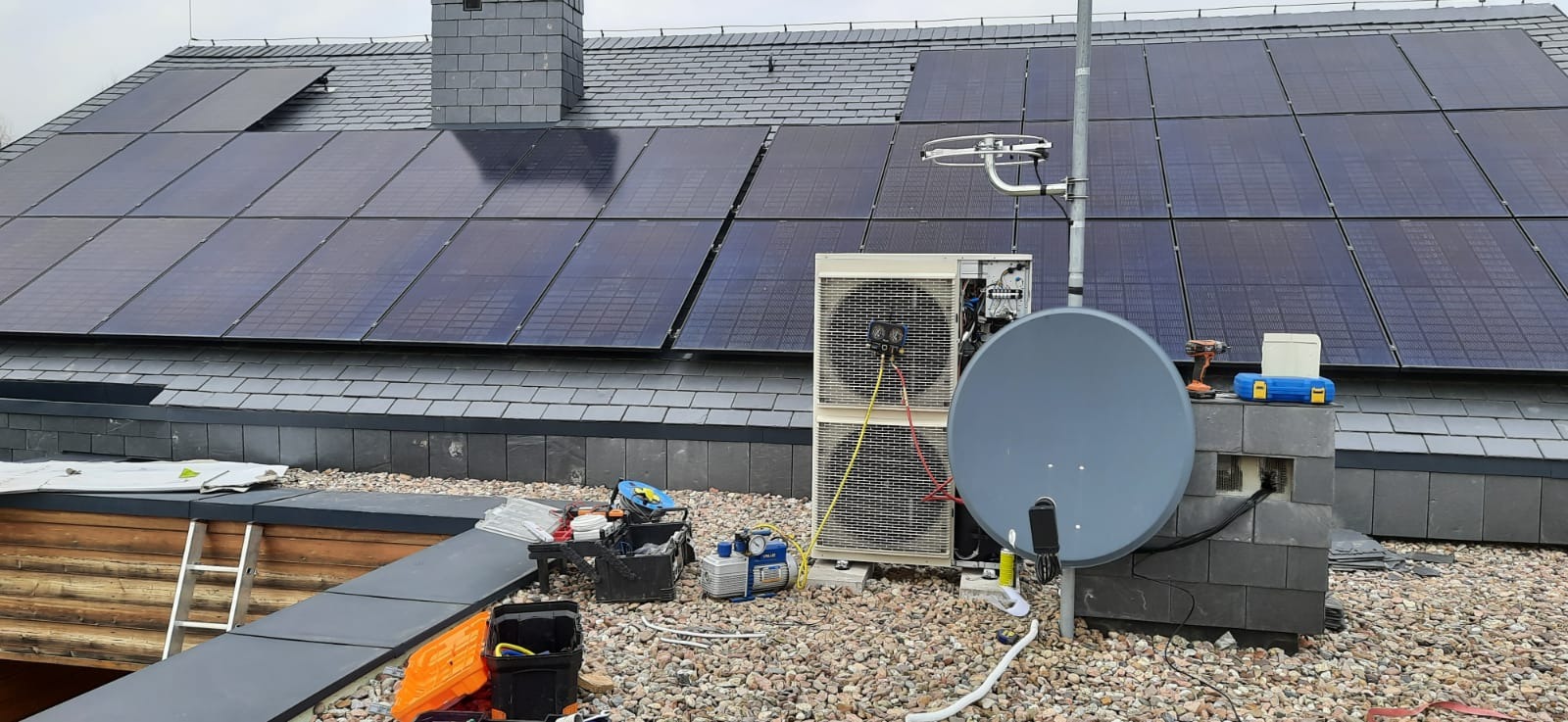Instalacja paneli fotowoltaicznych na dachu z szarej dachówki łupkowej, obok zainstalowany klimatyzator zewnętrzny z podłączonymi przewodami serwisowymi, widoczny również osprzęt monterski i antena...