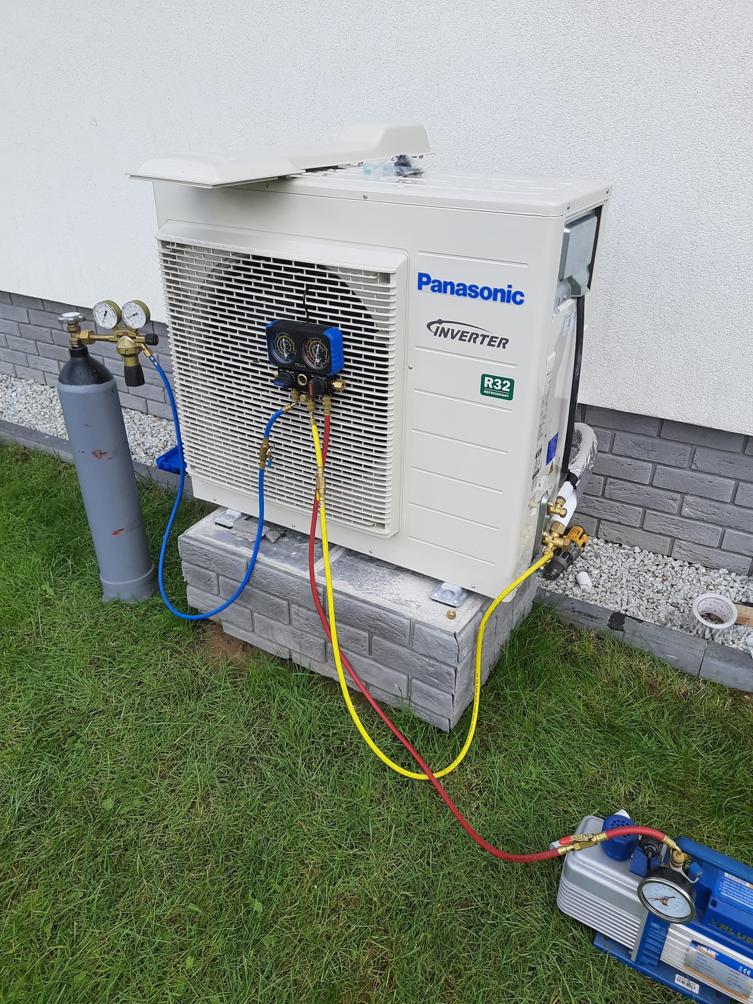 Biała jednostka zewnętrzna klimatyzacji Panasonic Inverter R32, umieszczona na szarej betonowej podstawie, podłączona do butli z gazem i pompy próżniowej za pomocą węży z manometrami podczas...