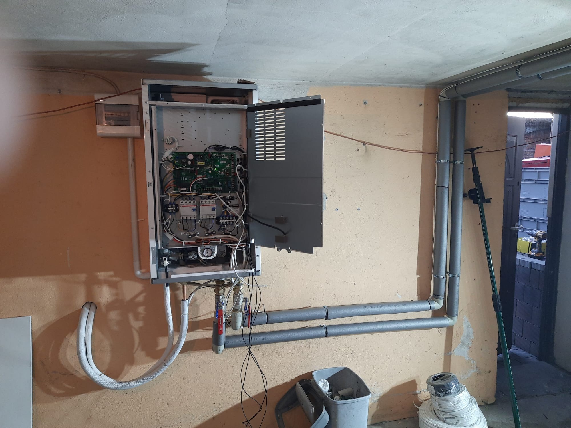 Otwarta obudowa urządzenia grzewczego z widocznymi elementami elektronicznymi i przewodami, rury instalacyjne prowadzone wzdłuż ściany, widoczny fragment pomieszczenia w stanie surowym.