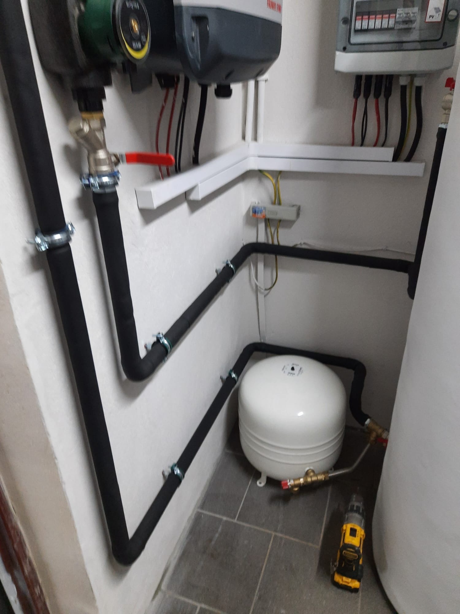 Instalacja z białym zbiornikiem wyrównawczym, czarnymi rurami izolowanymi i pompą obiegową, widoczne okablowanie elektryczne i skrzynka bezpiecznikowa na ścianie, w tle płytki podłogowe, w pobliżu...