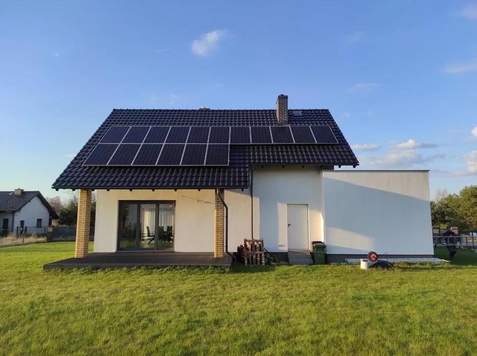 Biały dom jednorodzinny z ciemną dachówką i panelami słonecznymi na dachu, widok z trawnika w słoneczny dzień.