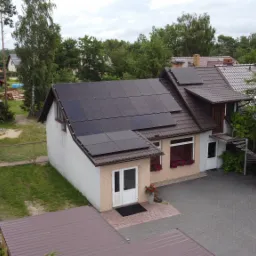 Dom jednorodzinny z brązowym dachem i panelami fotowoltaicznymi na dachu, widok z góry, kostka brukowa na podjeździe, kwiaty w doniczkach pod oknami.