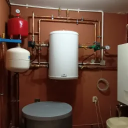 Instalacja z dwoma zbiornikami buforowymi, czerwonym i białym, oraz szarym zbiornikiem akumulacyjnym na tle brązowej ściany z miedzianymi rurami i zaworami.