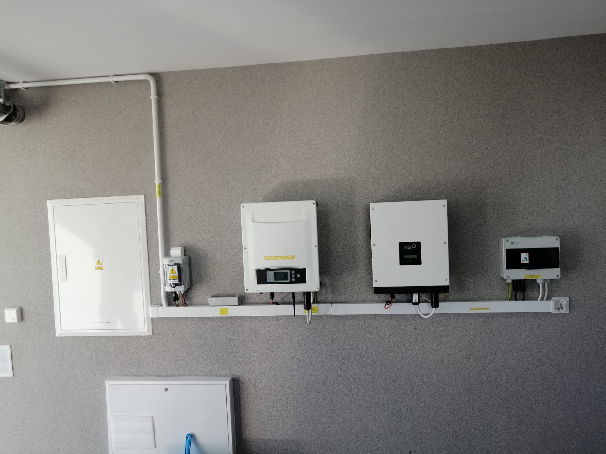 Dwa inwertery (Zeversolar i Fox) oraz skrzynka elektryczna zamontowane na ścianie z szarą tapetą strukturalną, widoczne okablowanie i rurki instalacyjne.