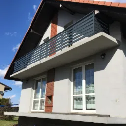 Szara balustrada balkonowa z poziomych profili na tle domu z czerwoną dachówką i błękitnym niebem.