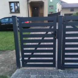 Nowoczesna, antracytowa brama wjazdowa z poziomymi szczeblami, widok na posesję z kostką brukową i domem w tle.