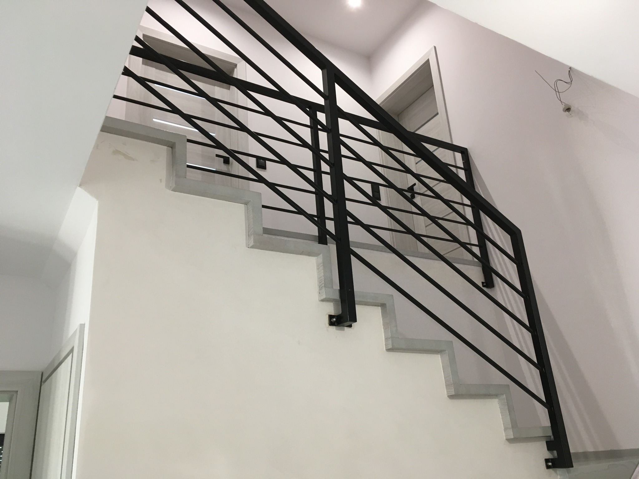 Nowoczesna klatka schodowa z metalową balustradą w kolorze czarnym, minimalistyczny design, białe ściany i szare stopnie, widoczne fragmenty drzwi.