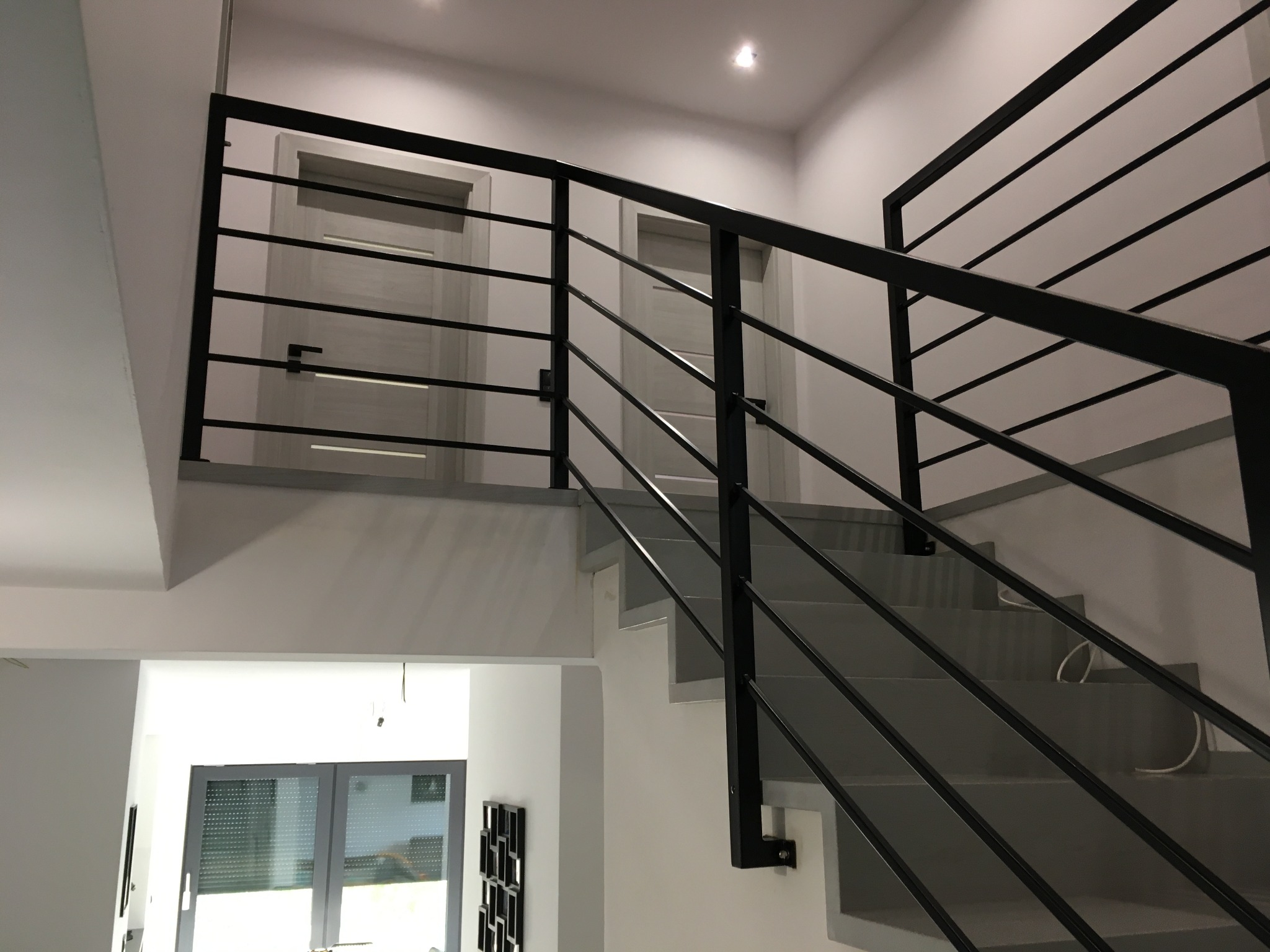 Nowoczesne schody z szarobetowymi stopniami i czarną metalową balustradą z poziomymi prętami, widok z dołu na górę, z widocznymi drzwiami w tle.