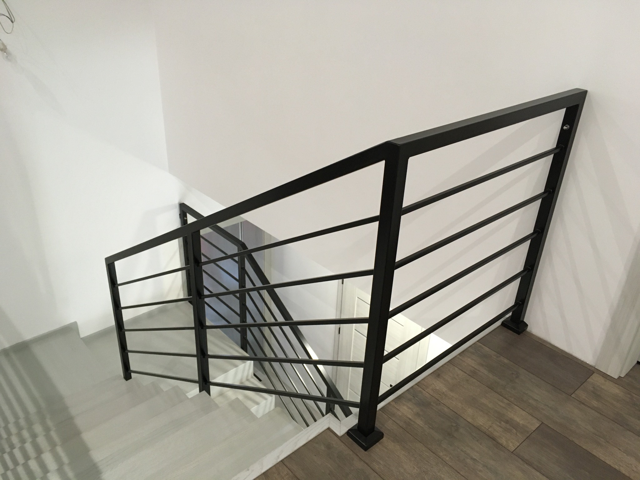 Nowoczesna, czarna metalowa balustrada schodowa z poziomymi prętami na białych schodach i drewnianej podłodze, widok z góry.
