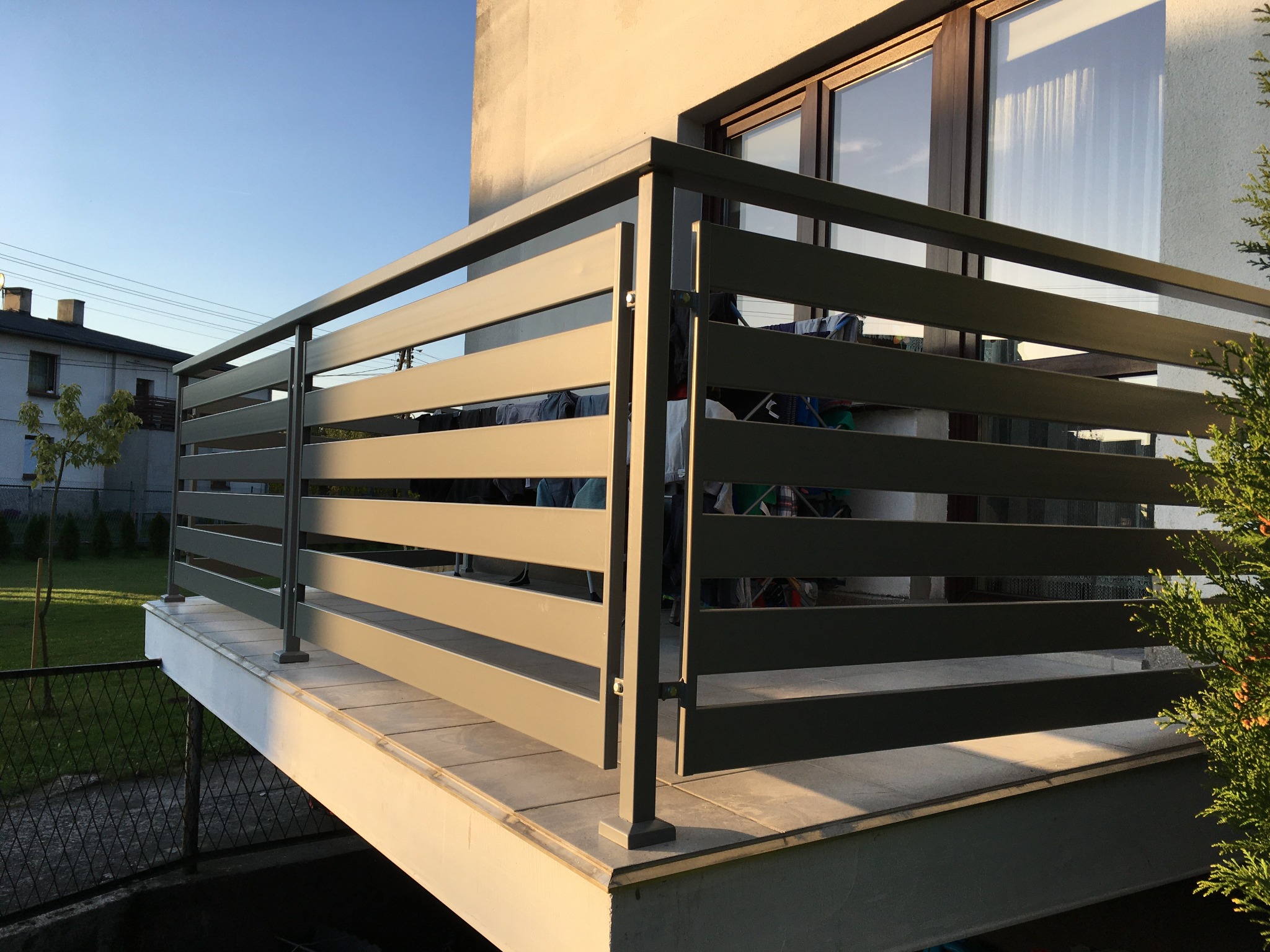 Nowoczesna balustrada balkonowa z poziomych, metalowych profili w kolorze szarym, zamontowana na płytkach, z widokiem na budynek mieszkalny i fragment ogrodu.