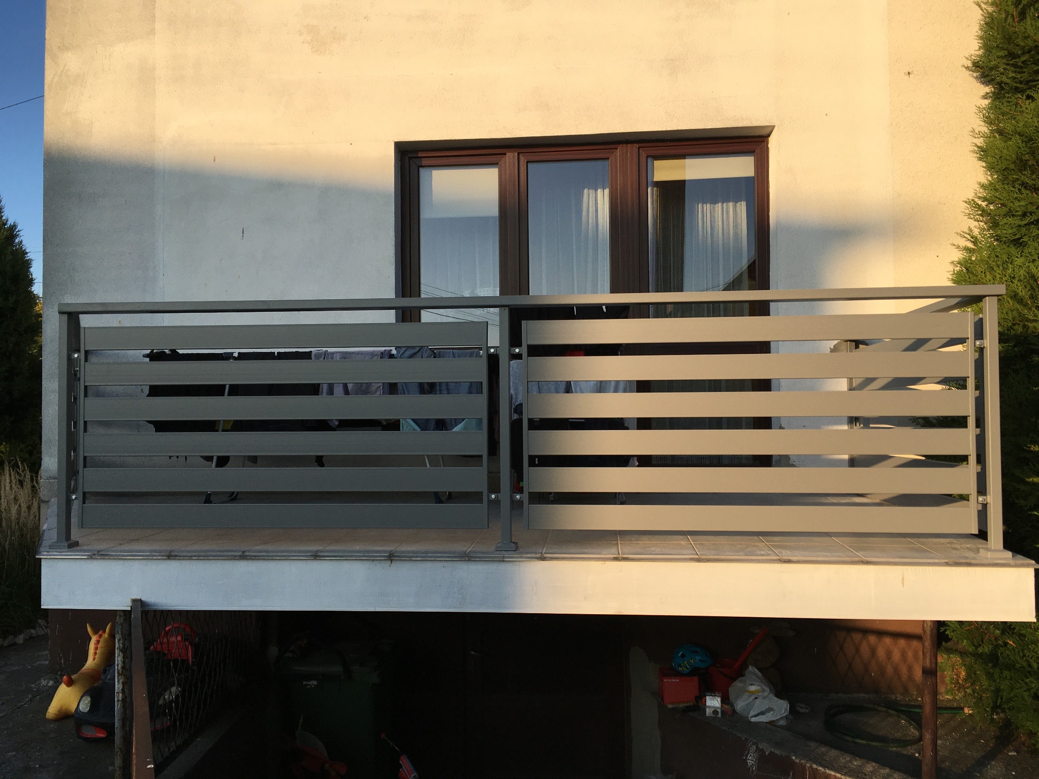 Szara, pozioma balustrada balkonowa z metalu, widok z dołu, z widocznym oknem i fragmentem elewacji budynku w tle, oraz elementami wyposażenia piwnicy pod balkonem.