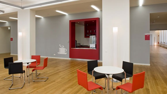 Jasne, przestronne wnętrze biurowe z parkietem, stolikami kawowymi z czerwonymi i czarnymi krzesłami, oraz czerwonym barem z ekspresem do kawy i lodówką na napoje, ozdobione naklejką filiżanki kawy...