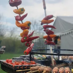 Przygotowanie potraw na grillu: szaszłyki z mięsa na patyku i kolorowe papryki zawieszone nad paleniskiem, w tle biały namiot.