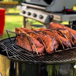 Żeberka BBQ w metalowym stojaku na czarnym grillu węglowym na zewnątrz, z drugim grillem widocznym w tle.