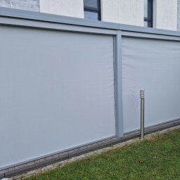 GK Katarzyna Górna - Dwa panele przesuwne z szarej tkaniny w metalowej ramie, tworzące osłonę tarasu, widoczne z zewnątrz, z lampą ogrodową obok.