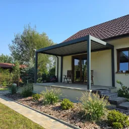 Nowoczesna pergola tarasowa z szarymi słupami i zadaszeniem lamelowym przy domu jednorodzinnym, widok na ogród z ozdobnymi trawami i ścieżką z kostki brukowej.