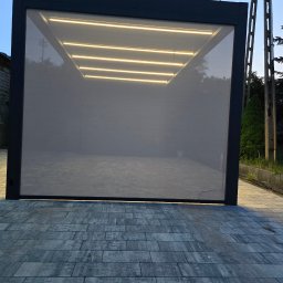 GK Katarzyna Górna - Nowoczesna pergola z szarą tkaniną osłonową i oświetleniem LED, ułożona na kostce brukowej w odcieniach szarości.