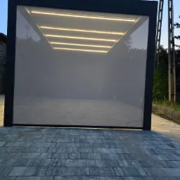 Nowoczesna pergola z szarą tkaniną osłonową i oświetleniem LED, ułożona na kostce brukowej w odcieniach szarości.