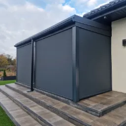Zewnętrzna zabudowa tarasu z grafitowymi roletami pionowymi, widok z boku na schody wejściowe i fragment ogrodu.