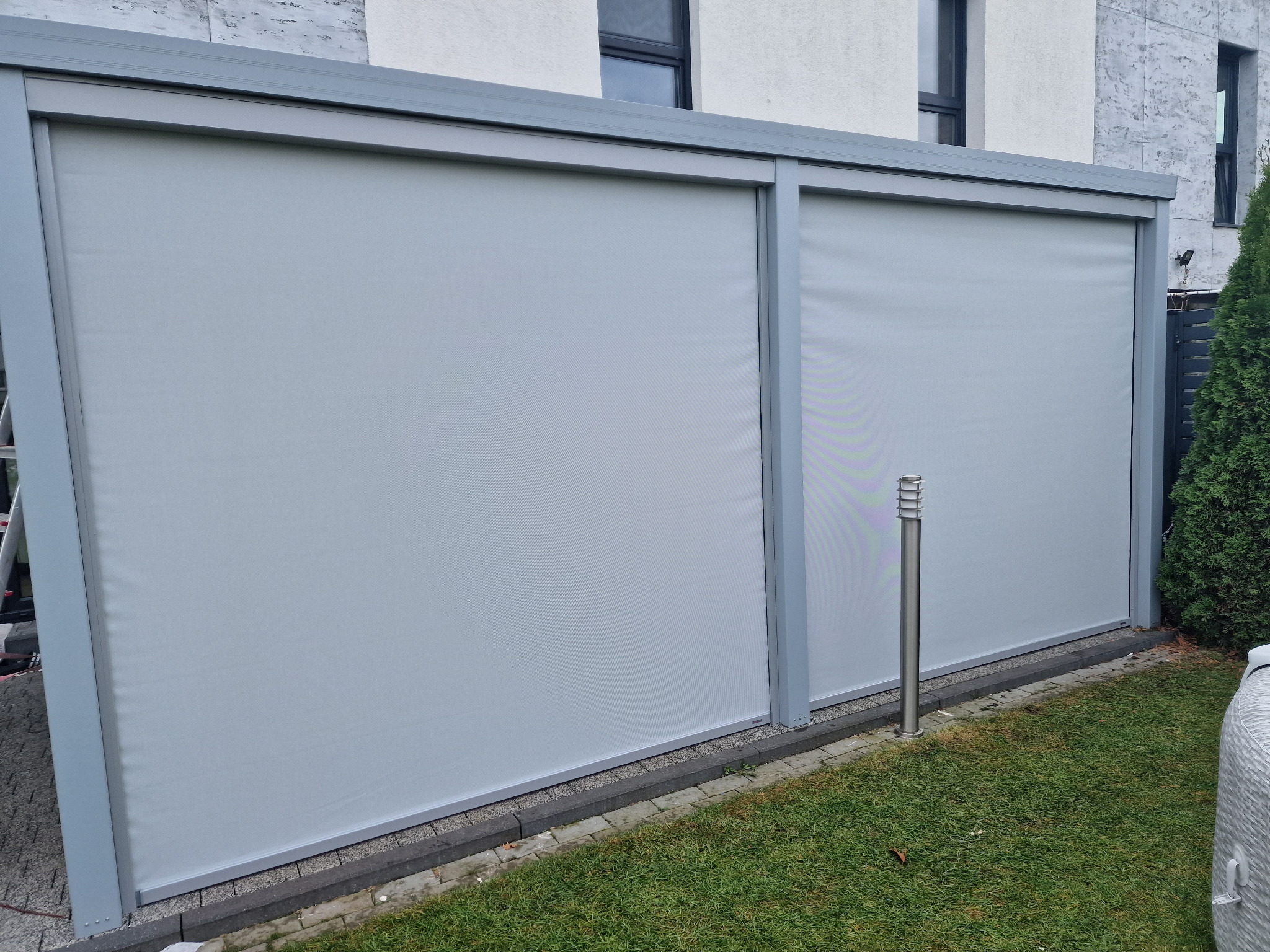 Dwa panele przesuwne z szarej tkaniny w metalowej ramie, tworzące osłonę tarasu, widoczne z zewnątrz, z lampą ogrodową obok.