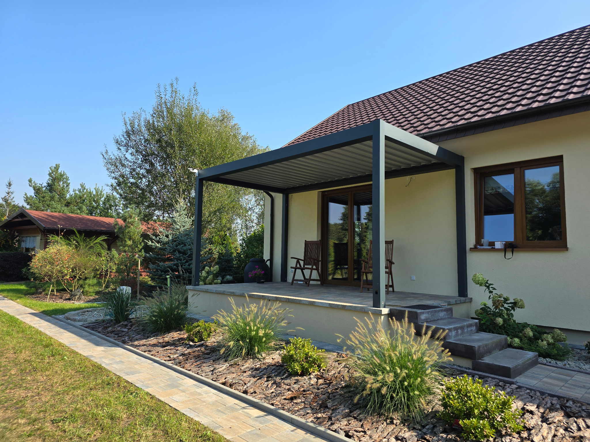 Nowoczesna pergola tarasowa z szarymi słupami i zadaszeniem lamelowym przy domu jednorodzinnym, widok na ogród z ozdobnymi trawami i ścieżką z kostki brukowej.