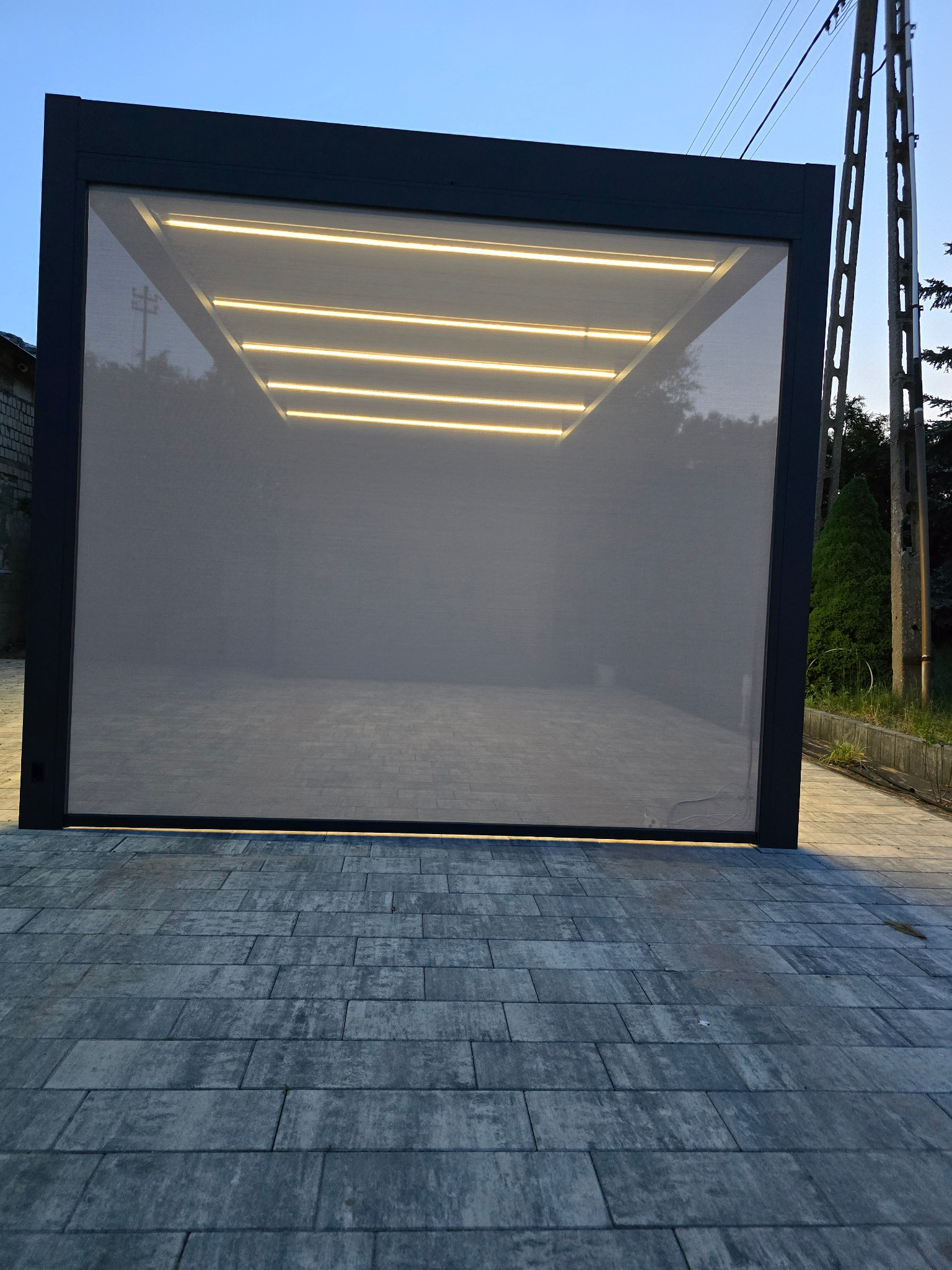 Nowoczesna pergola z szarą tkaniną osłonową i oświetleniem LED, ułożona na kostce brukowej w odcieniach szarości.