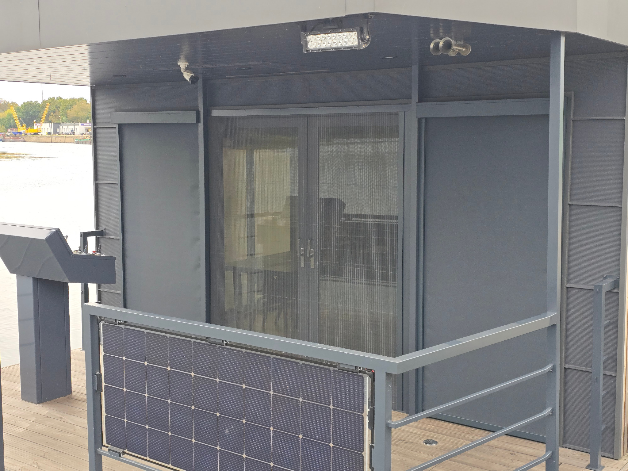 Szare, nowoczesne zabudowy balkonu z panelami słonecznymi i moskitierą plisowaną w drzwiach balkonowych, widok na rzekę i dźwigi w tle.